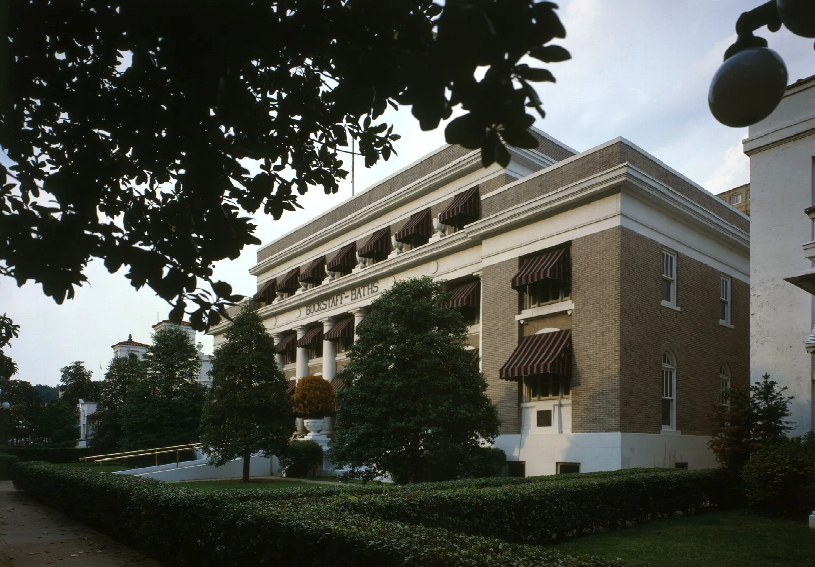 The front of a classical brick and stucco bathhouse behind trimmed hedges