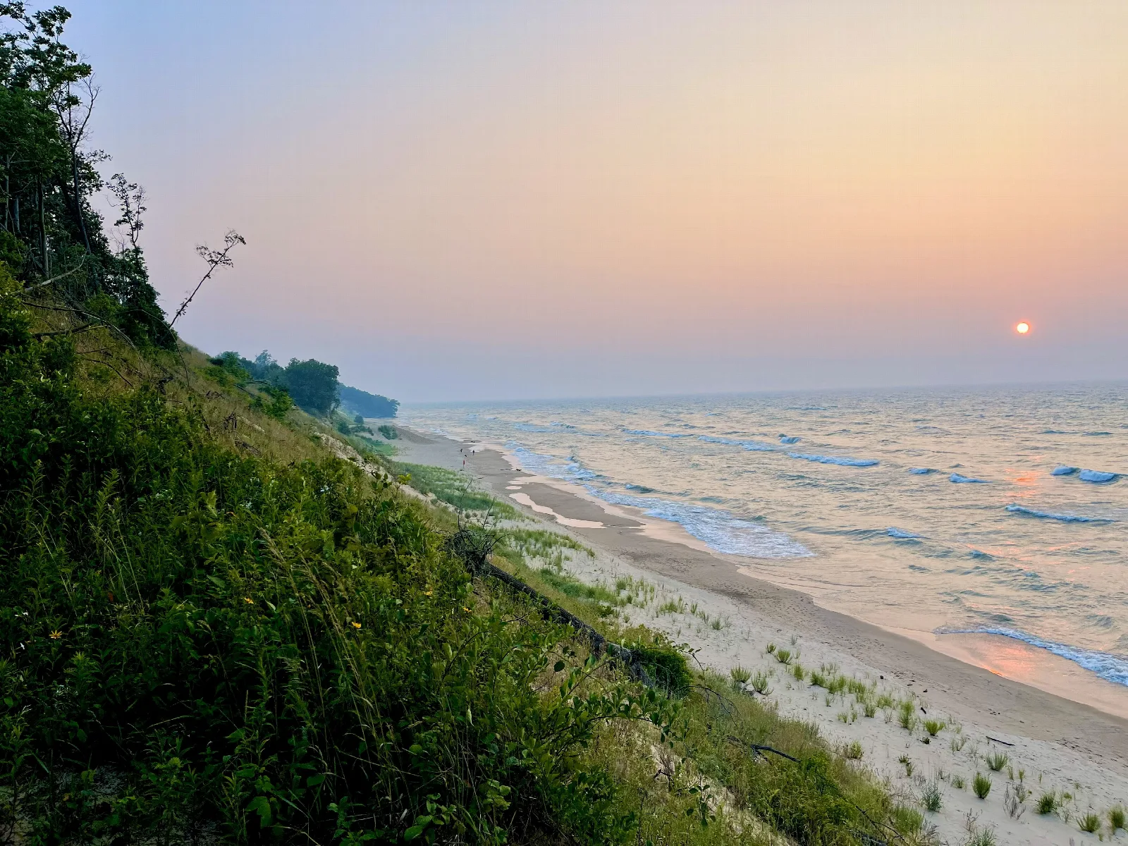 A green dune bluff dropping to a long sandy beach and Lake Michigan at sunset