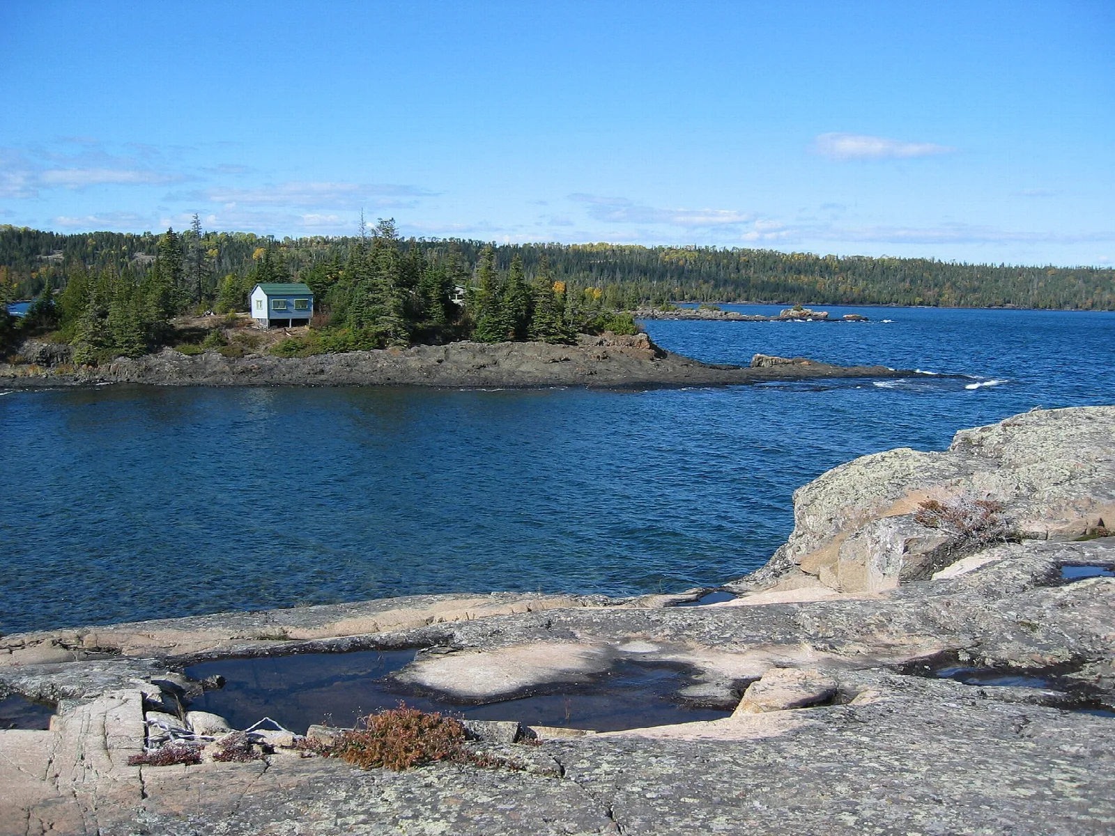 A rocky shoreline looking across a bay to a small cabin on a forested point