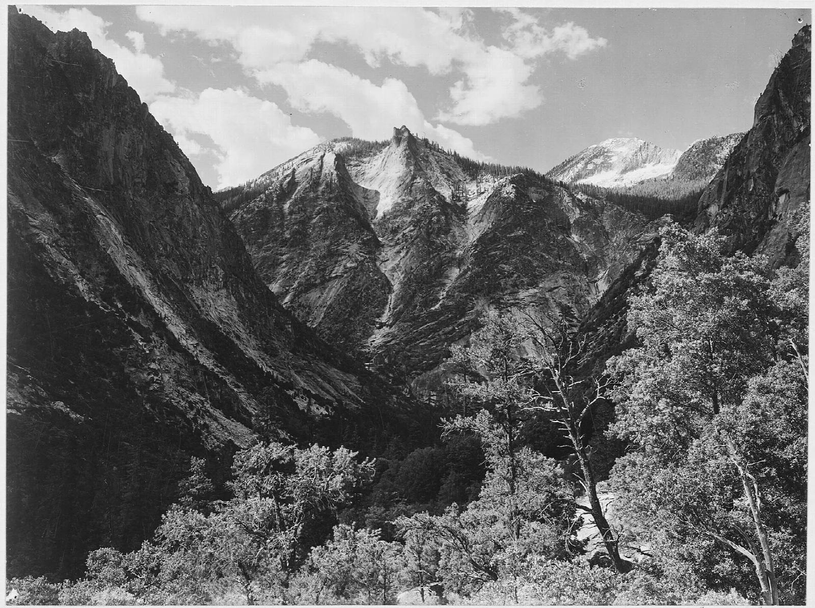 Black-and-white photograph of a forested valley beneath jagged granite peaks