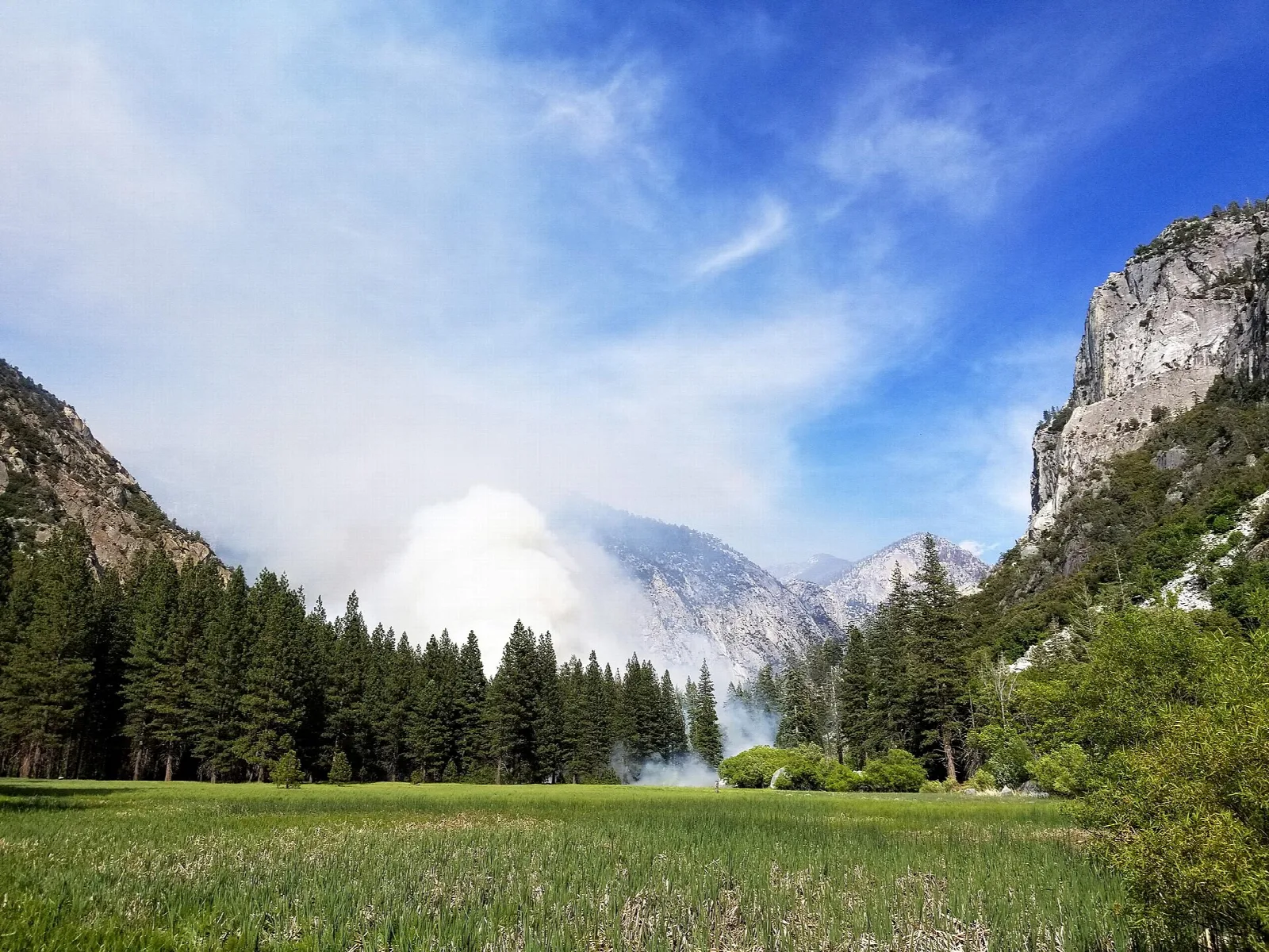 A green meadow and pines below granite cliffs, with smoke from a prescribed burn drifting beyond