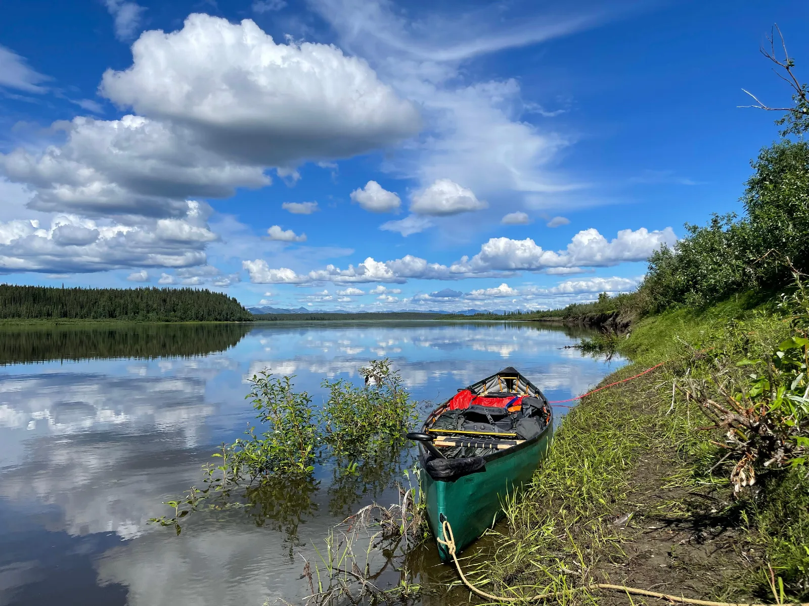 A green pack canoe sits on the bank of the Kobuk River. Clouds are mirrored on the water\'s surface.