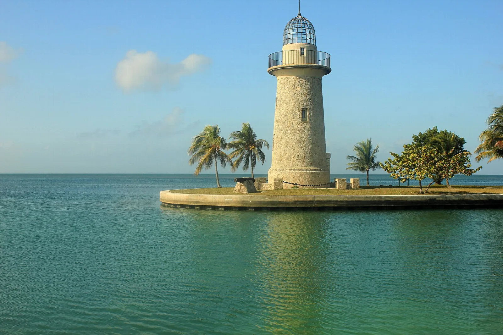 A white stone lighthouse on a seawall, flanked by palms, reflected in calm turquoise water