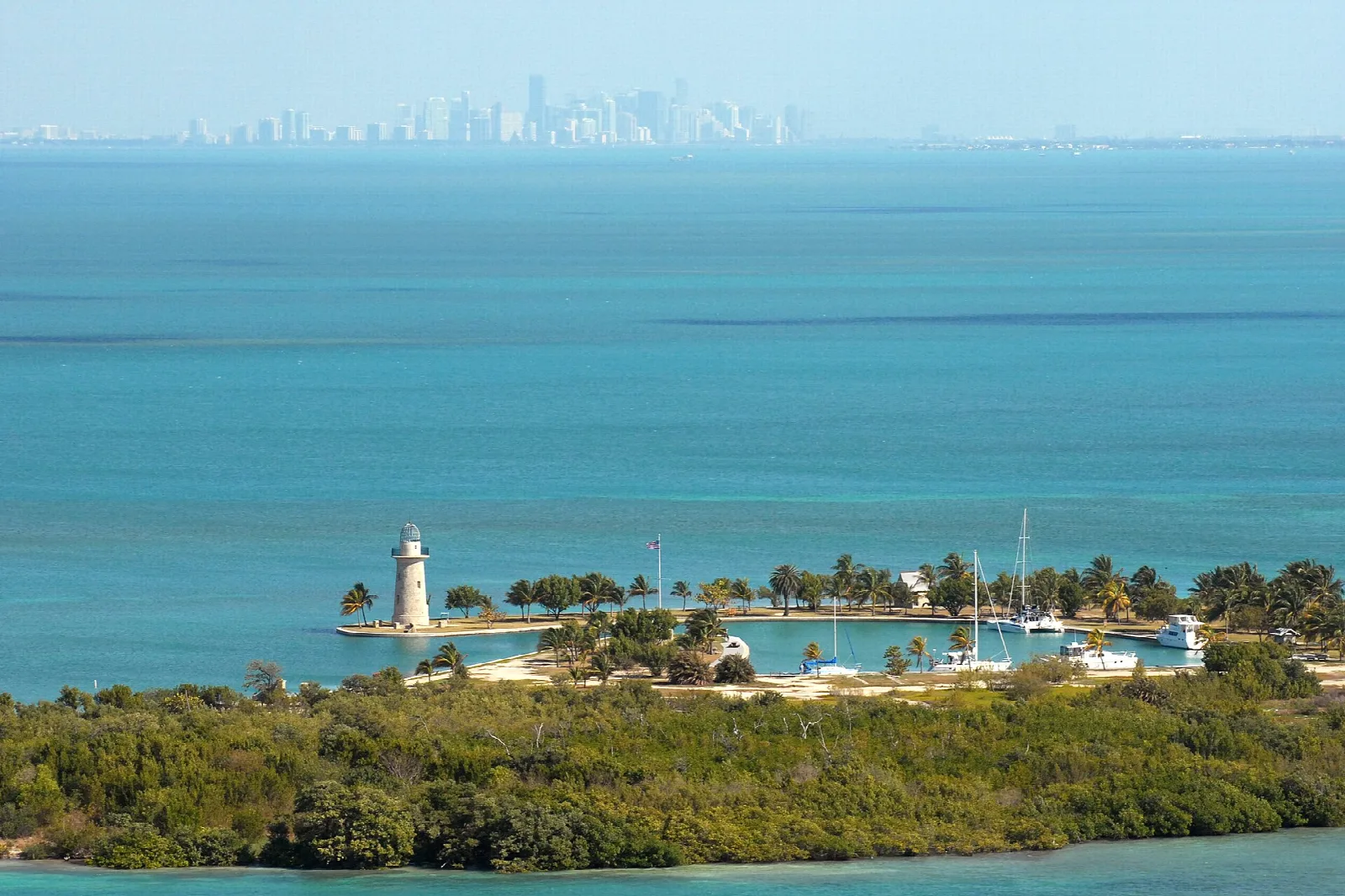 A small key with a lighthouse and harbor in a turquoise bay, the Miami skyline faint in the distance