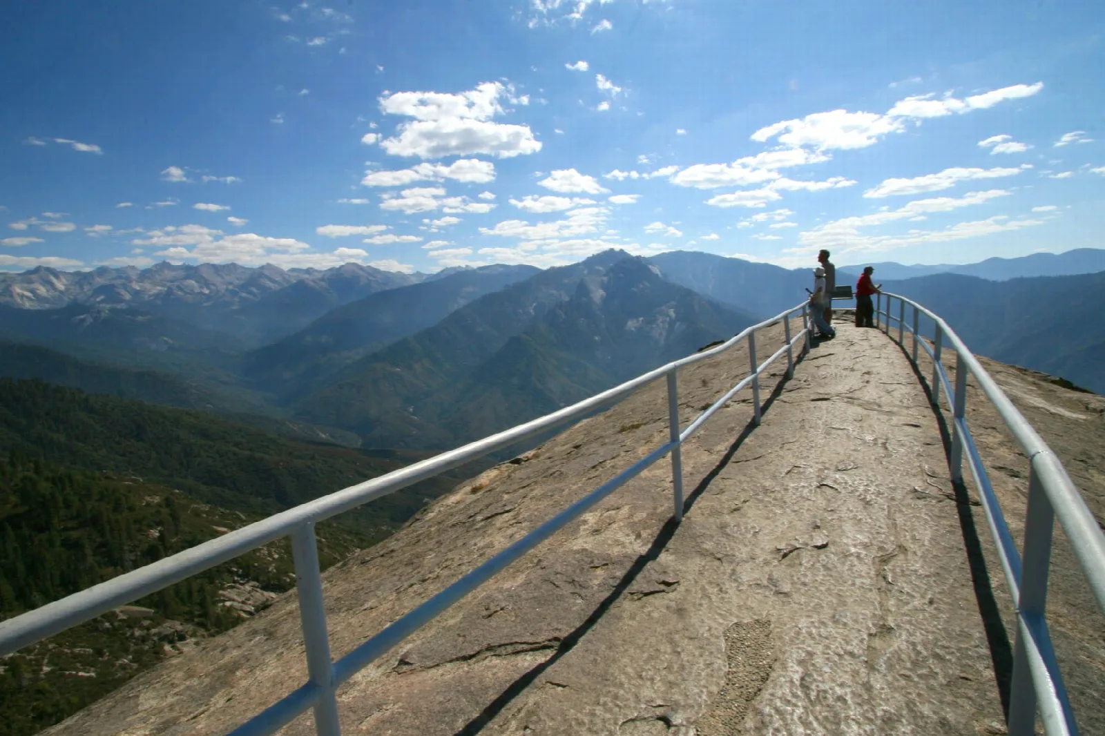 Hikers at a railed summit walkway on a granite dome above layered blue mountain ridges