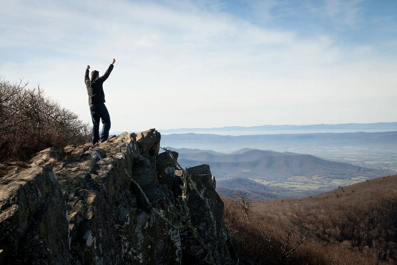 A hiker raising their arms on a rocky summit above hazy blue ridges