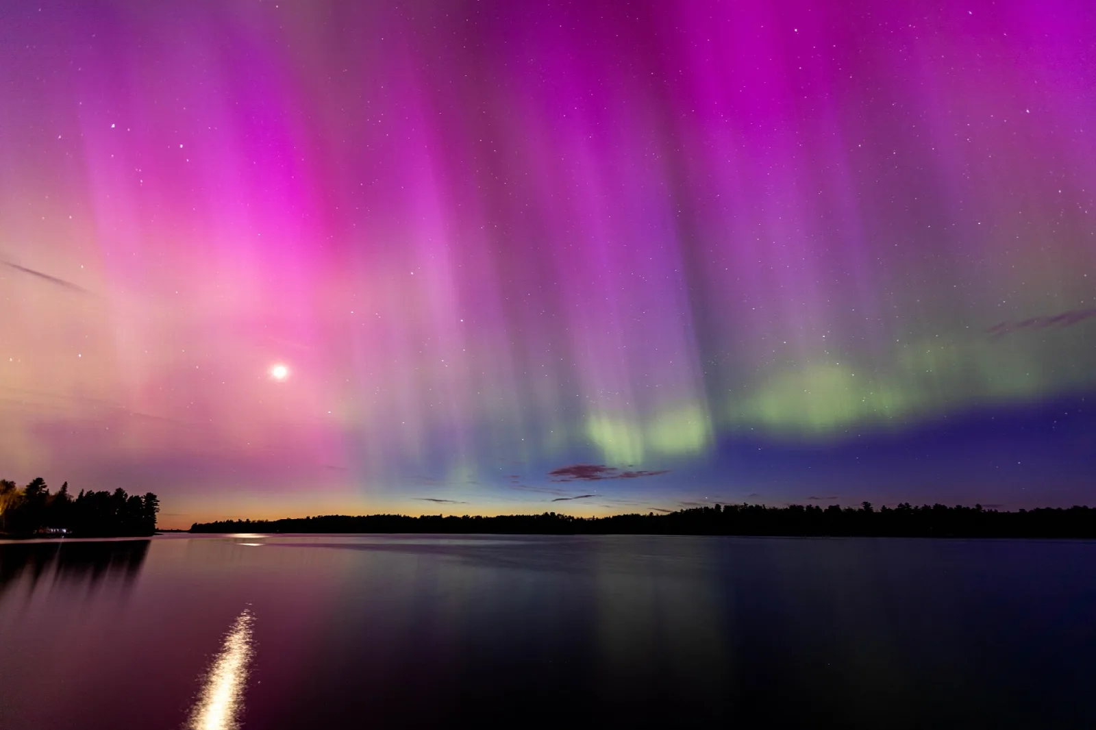 Pink and green aurora filling the night sky above a dark lake shore