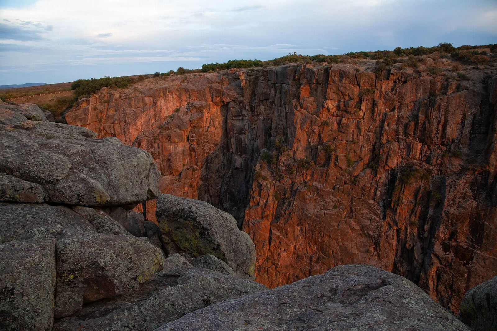 A steep canyon wall glowing red in the last light beyond granite boulders on the rim