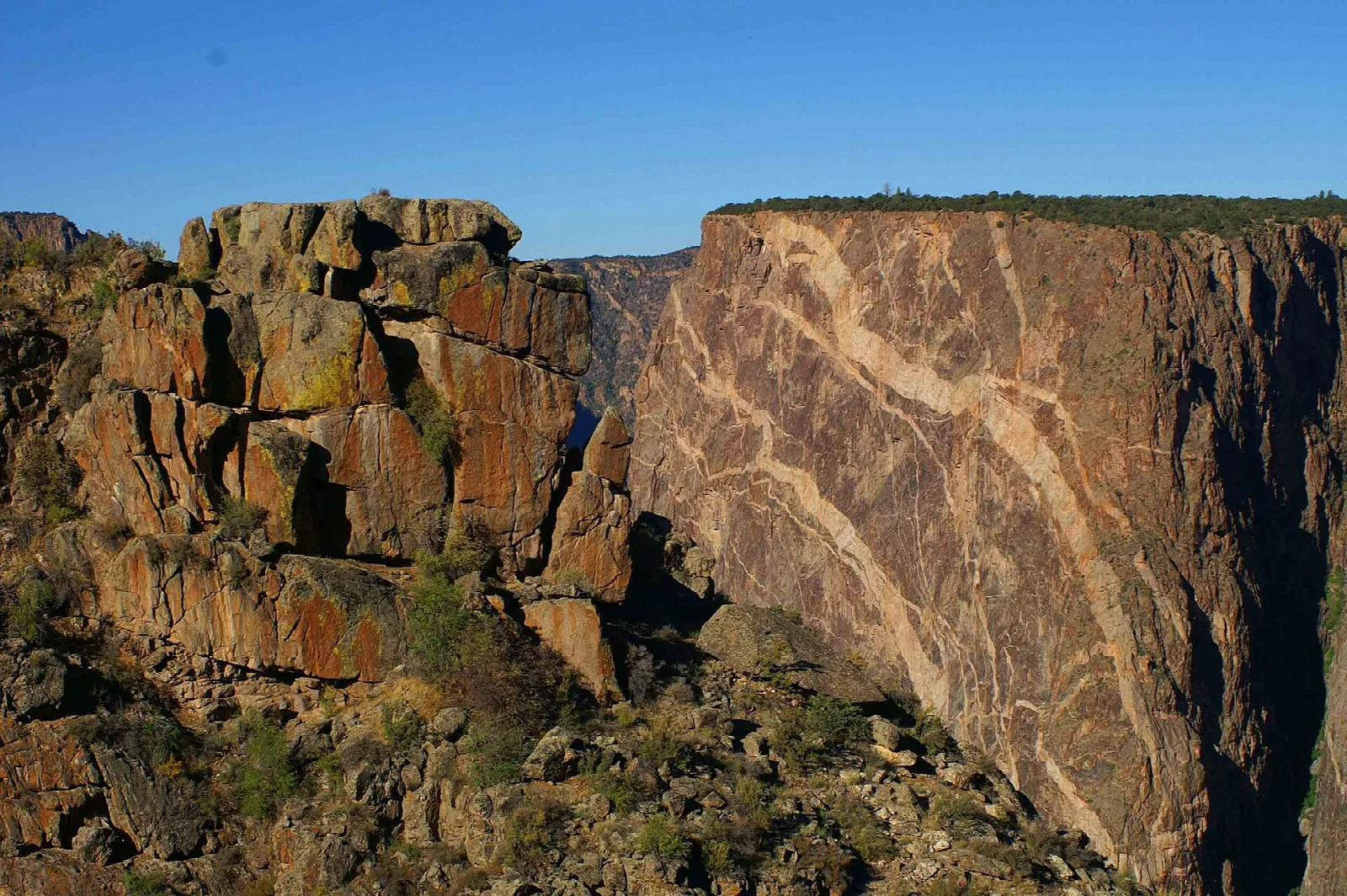 A sheer dark canyon wall streaked with pale pink veins of rock, lit by soft light under a blue sky