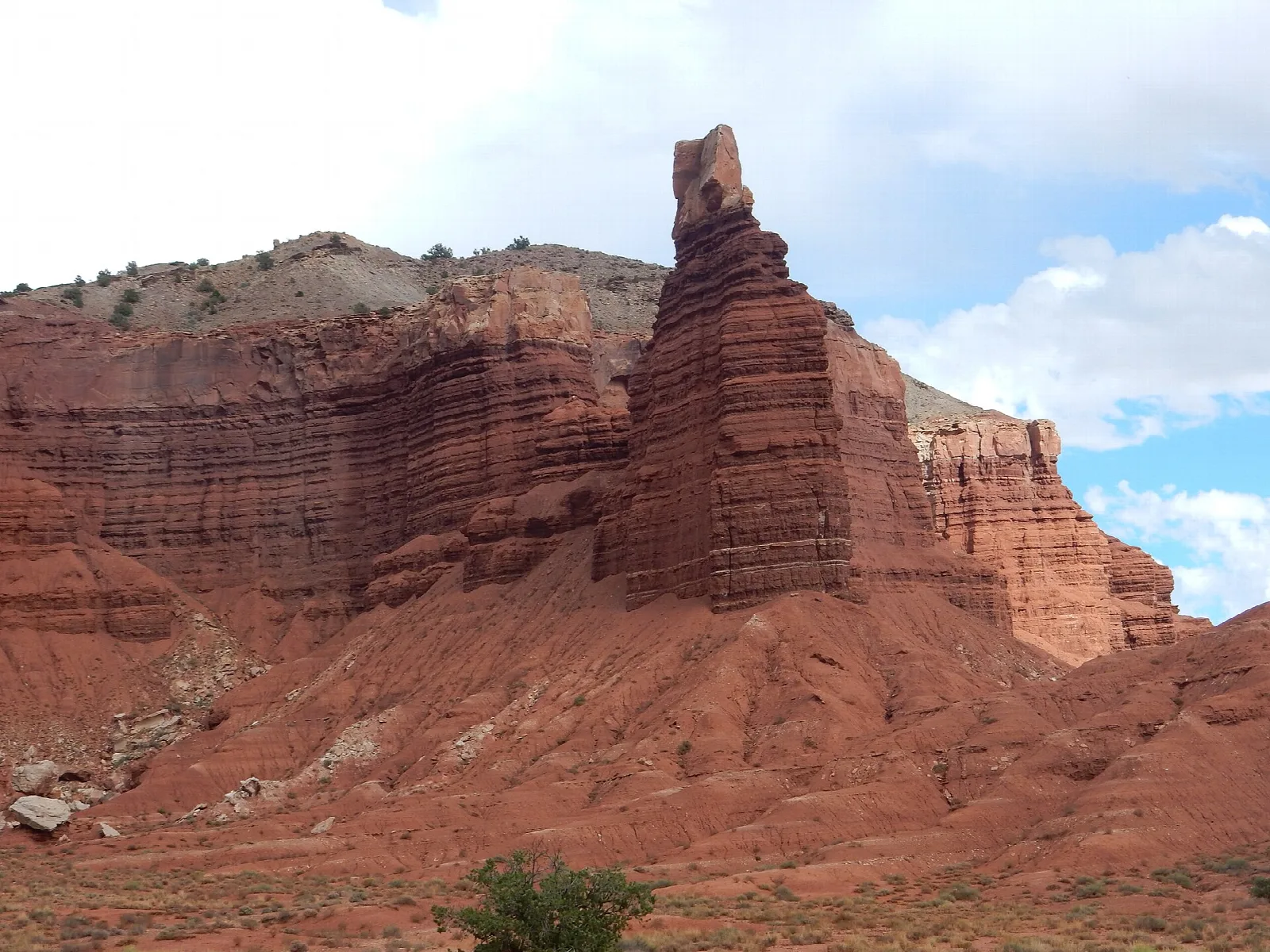 A dark red, layered rock spire on a rust-colored slope under scattered clouds