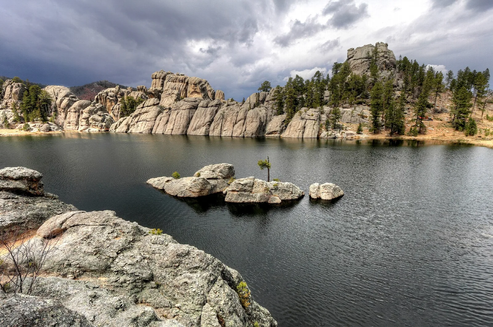Dark lake with granite boulders and a rocky ridge under stormy clouds