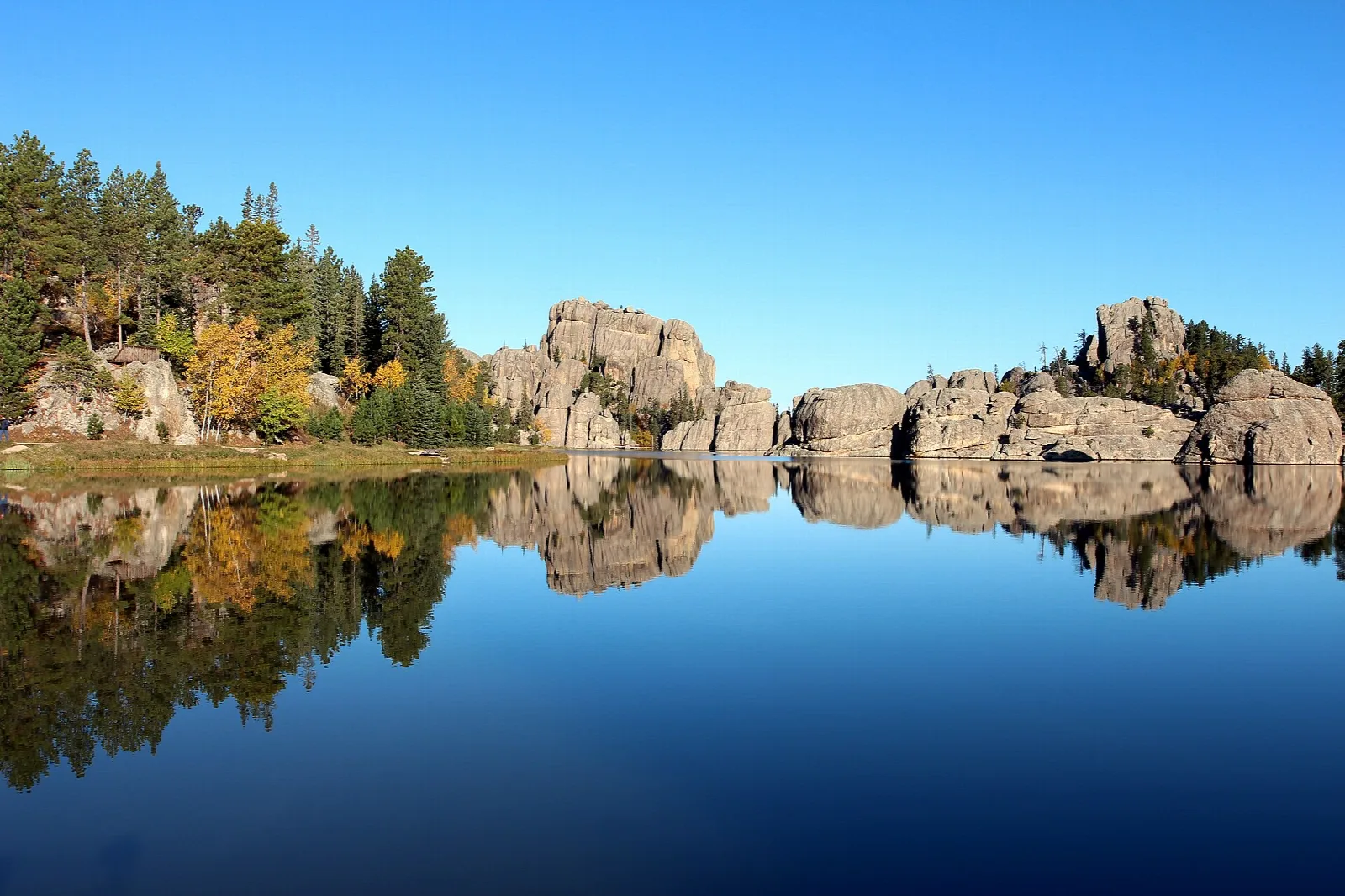 Calm lake reflecting rocky granite outcrops and pines under a blue sky