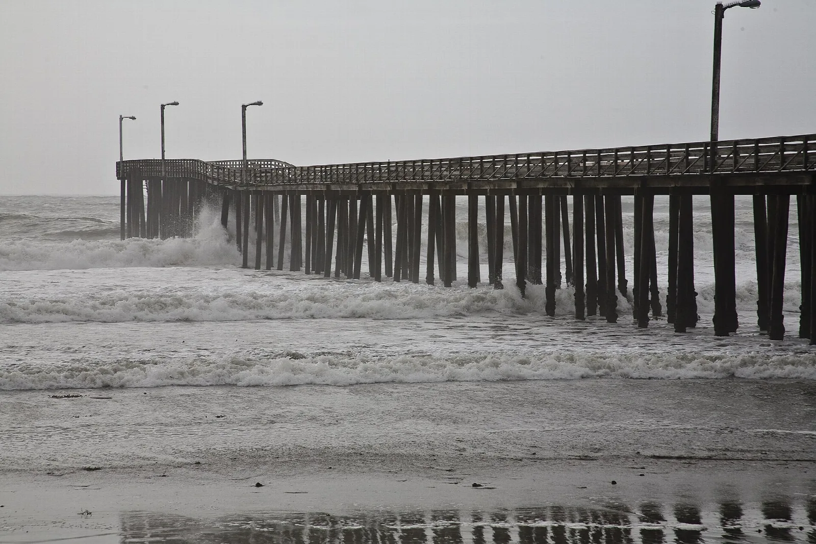 Waves breaking around a long wooden pier in black and white