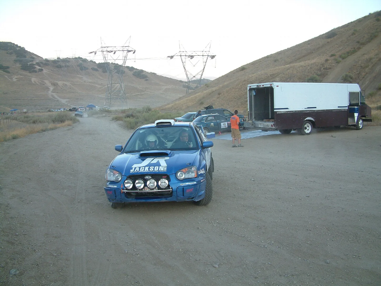Blue rally car on a dirt road with a trailer and power lines behind