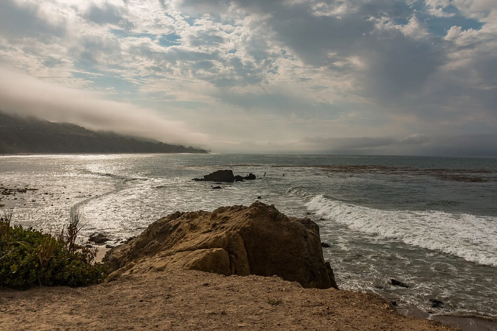 Rocky bluff above a surf-washed beach with a fog-topped ridge beyond