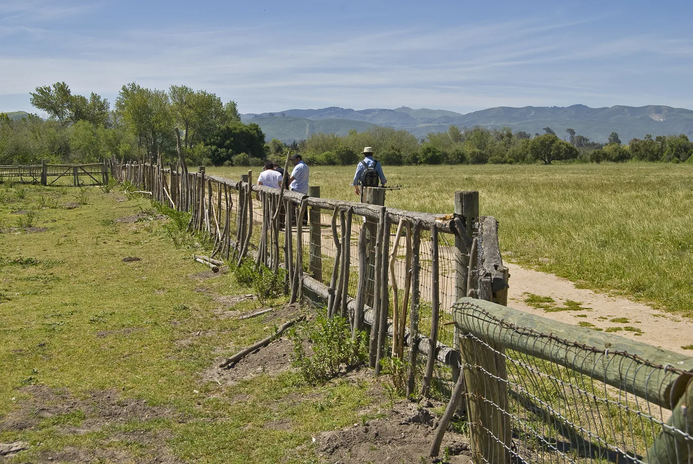 Wooden fence along a green meadow with hills in the distance and people walking