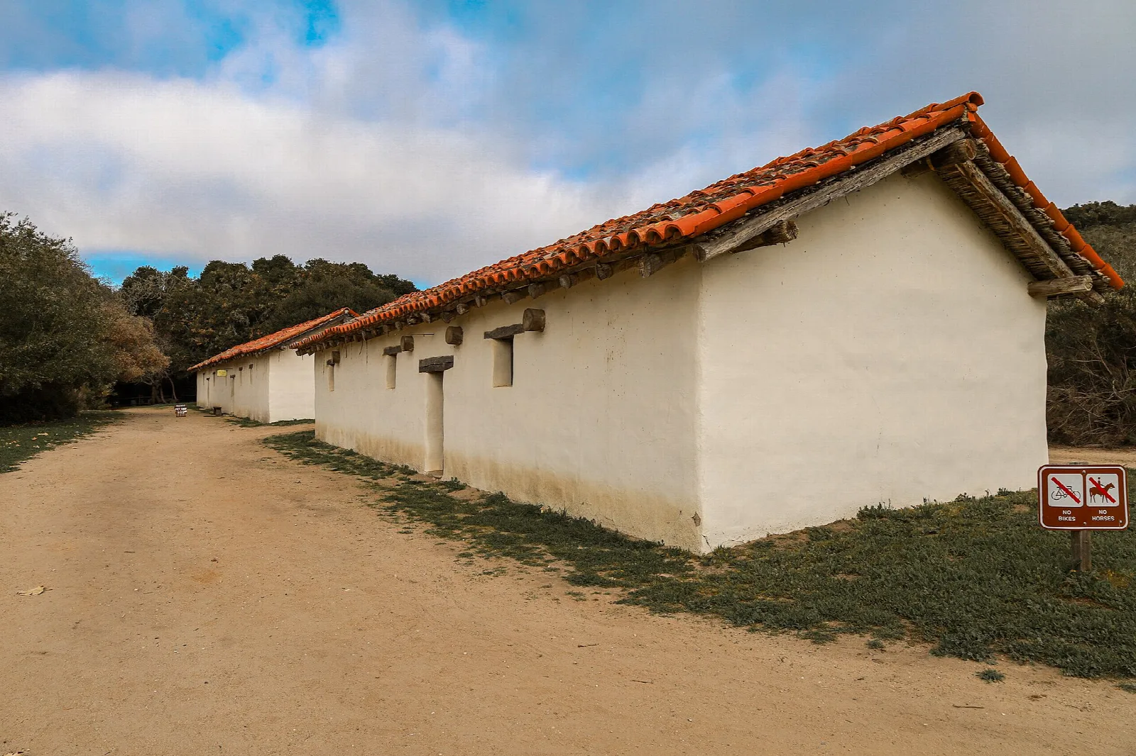 Long whitewashed adobe building with a red tile roof beside a dirt path