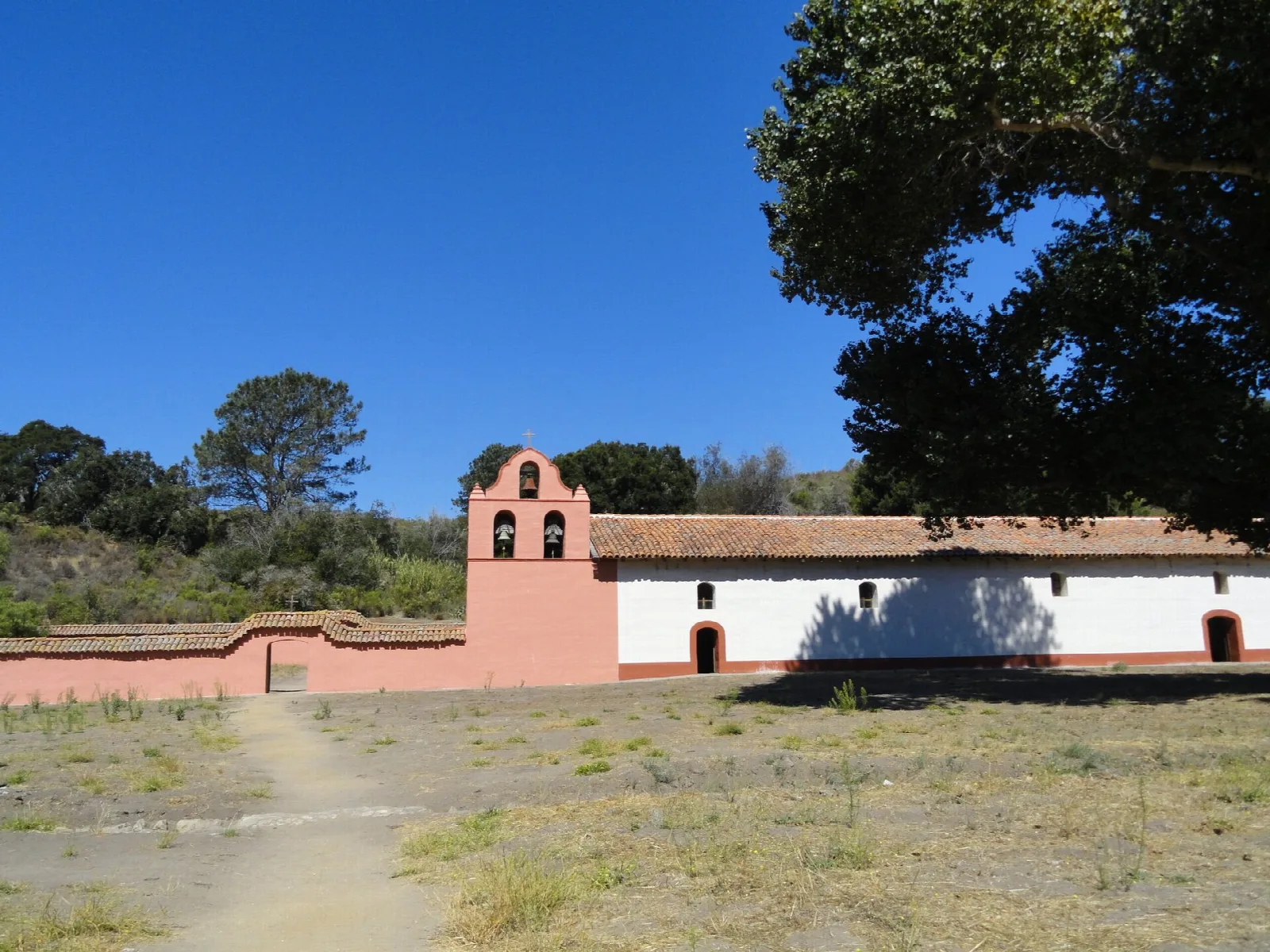 Pink-walled mission chapel with a bell gable beside an open dirt field and trees