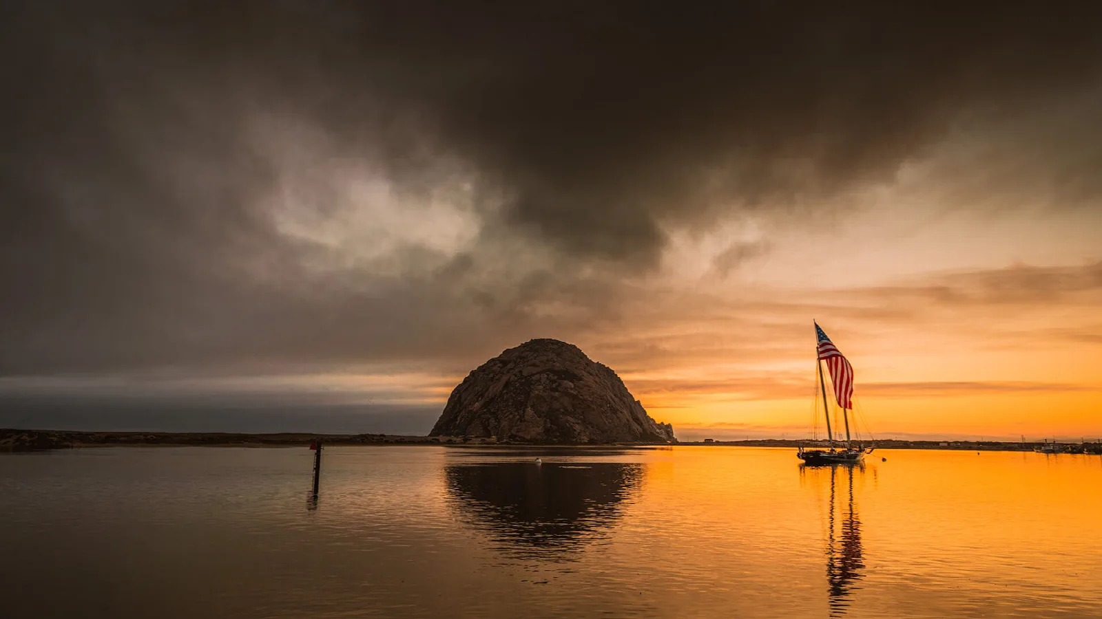 Morro Rock silhouetted on the water under a dramatic sunset sky with a flag