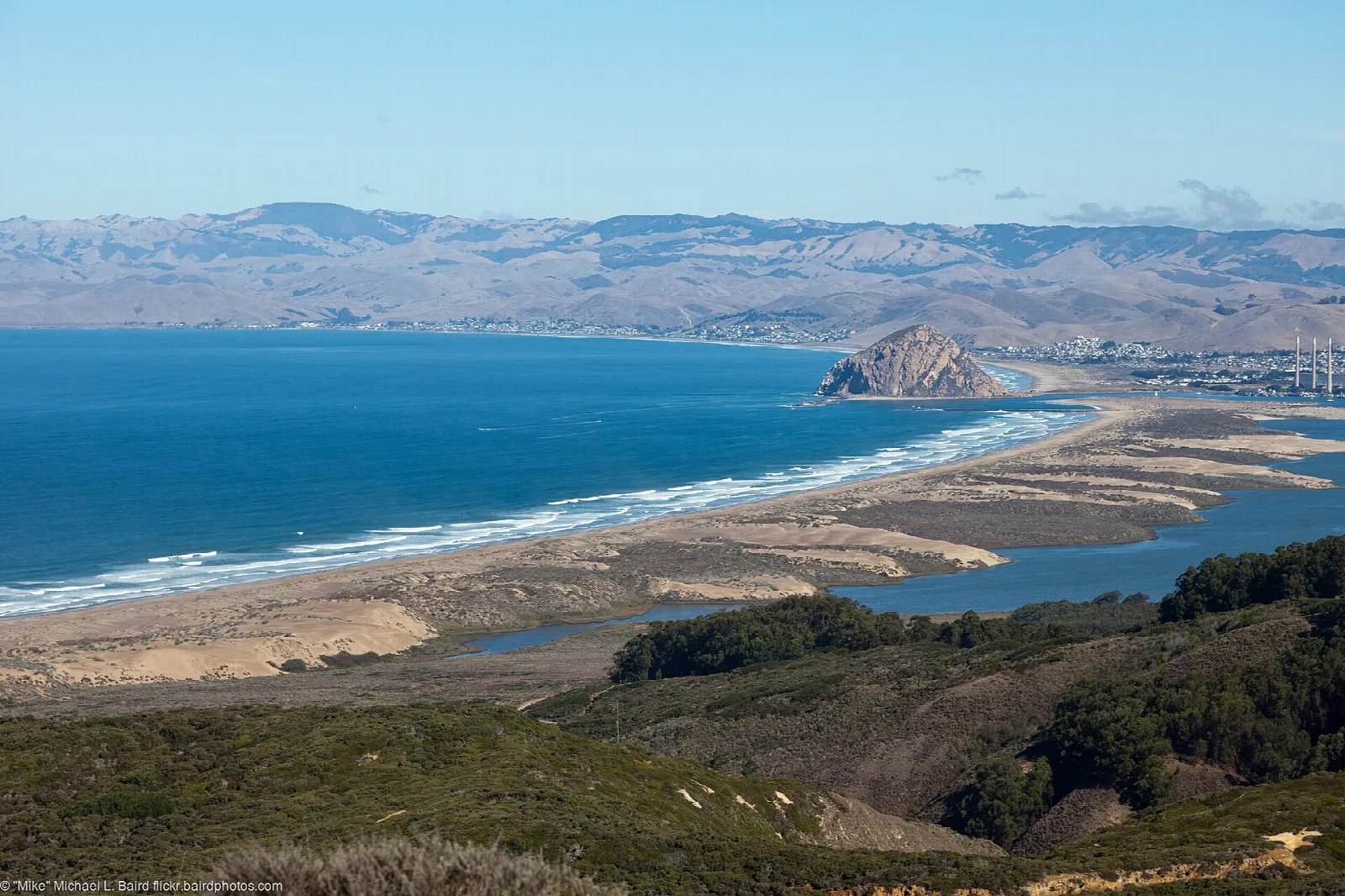 Coastline curving along blue water to a distant rock and sandspit, seen from a hilltop
