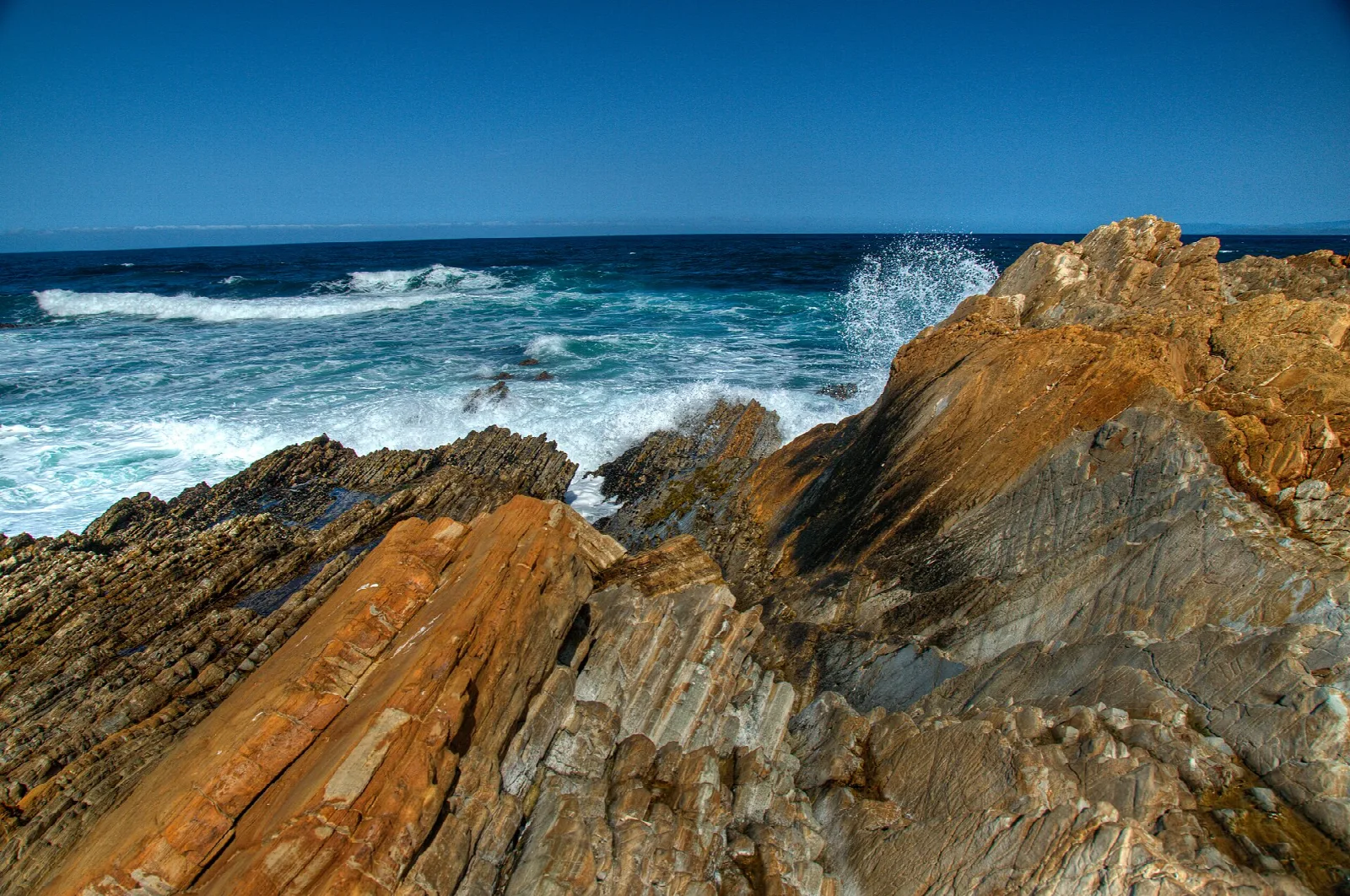 Layered orange and gray rock ledges above surf and deep blue sky
