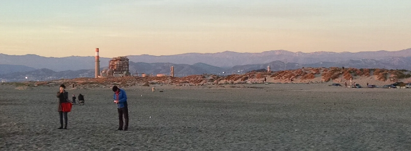 Wide flat beach with a few people at dusk and a distant power plant and mountains