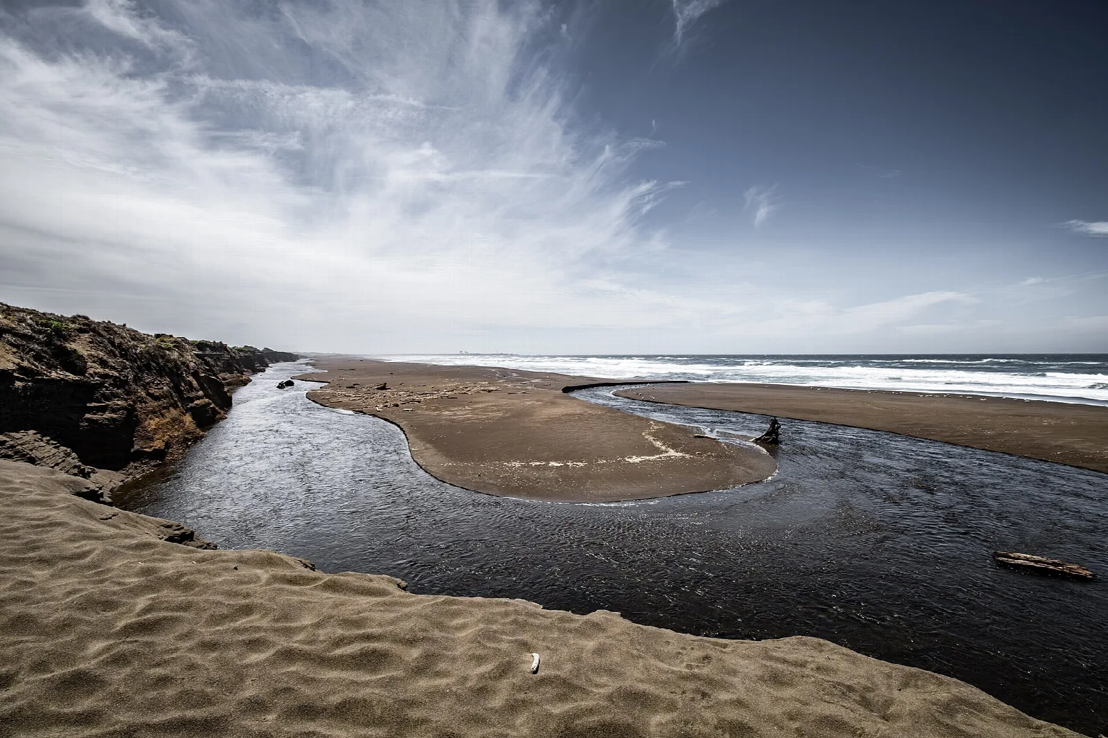 Creek winding through sand to the ocean under wispy clouds