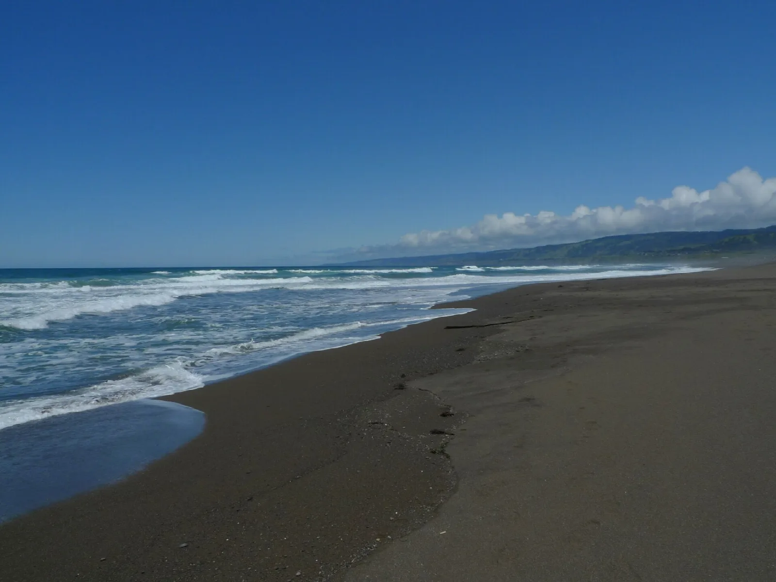 Dark sand beach with surf and a distant headland under blue sky