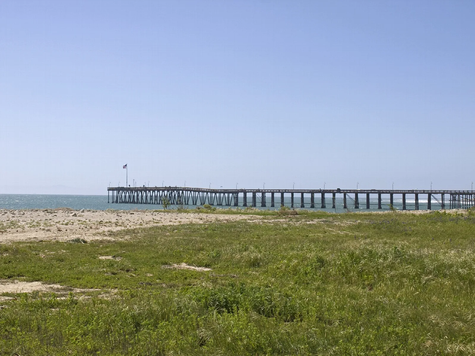 Long pier seen across grass and sand under a pale sky