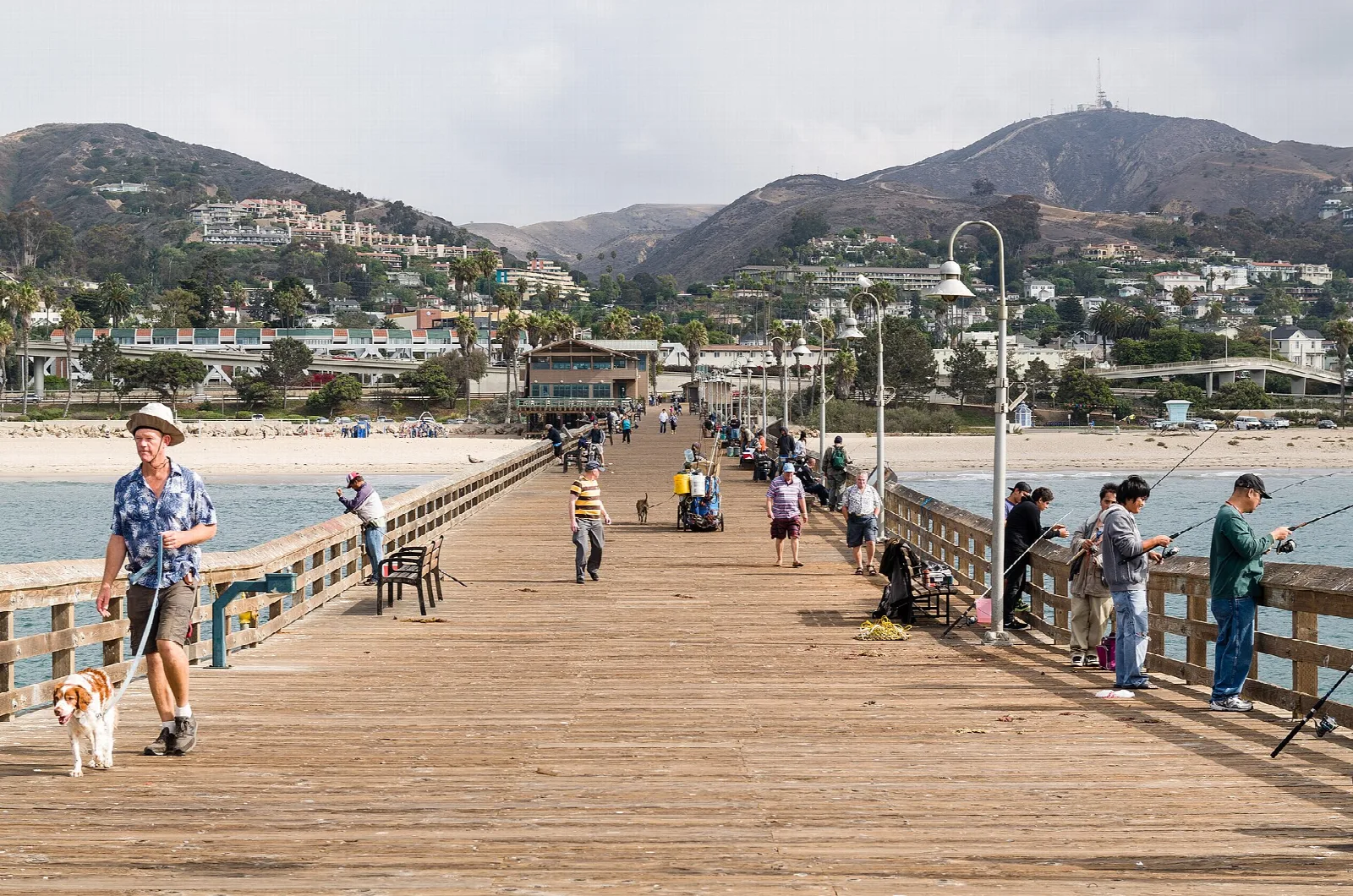 People walking and fishing on a wooden pier with hills behind