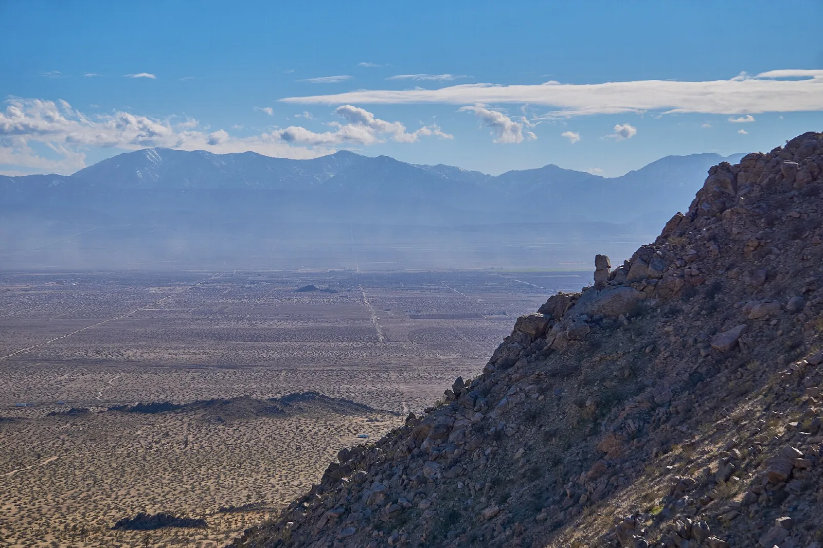 Rocky butte slope above a flat desert valley with mountains beyond