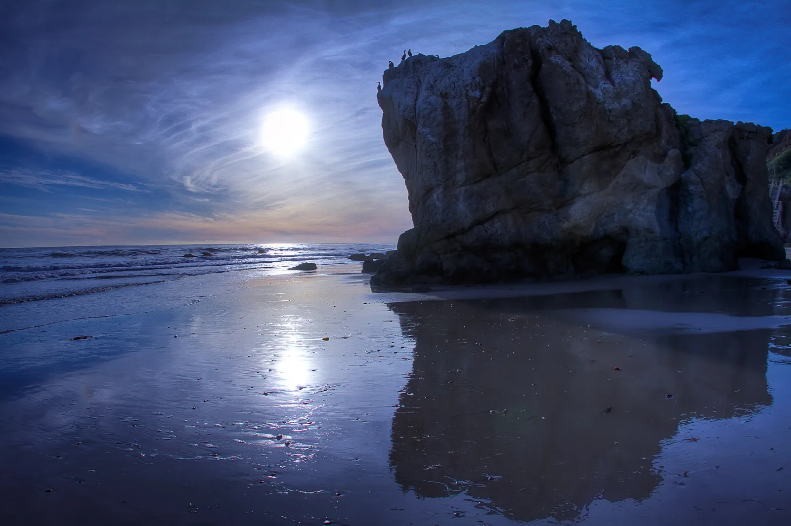 Large sea stack on a wet sandy beach with the sun low over the water