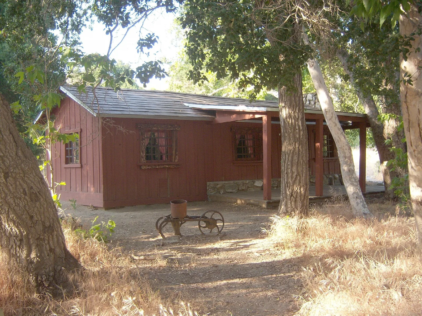 Small red wooden cabin shaded by trees, with an old plow in the yard