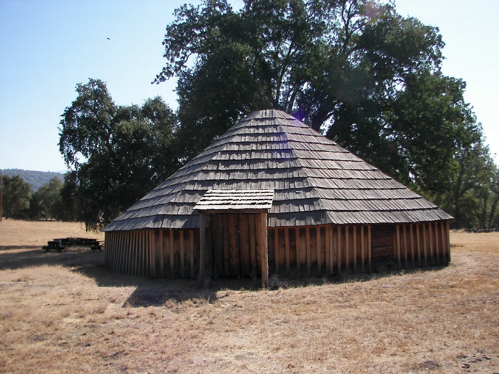 Round house with a shingled cone roof beneath large oaks