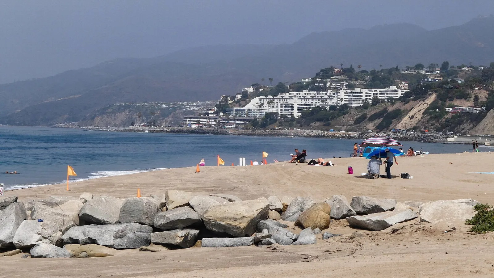 Beachgoers on sand beside a rock jetty with coastal hills behind