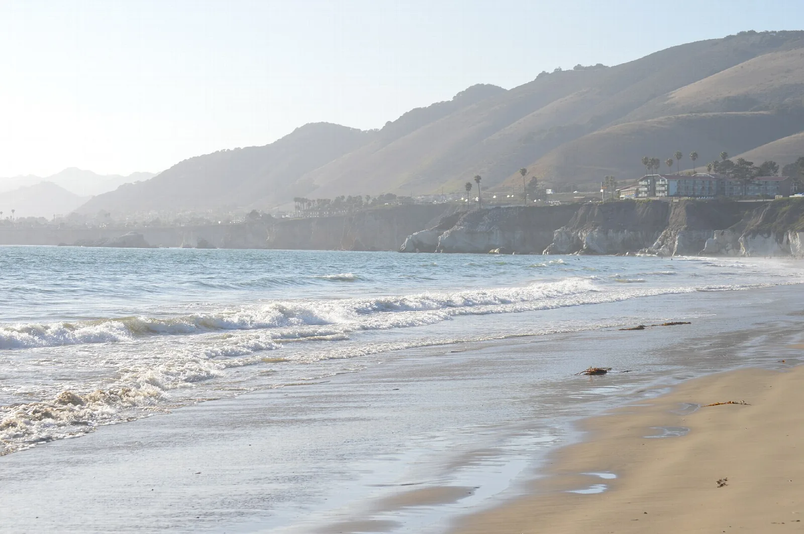 Surf rolling onto a sandy beach below pale coastal cliffs and hills