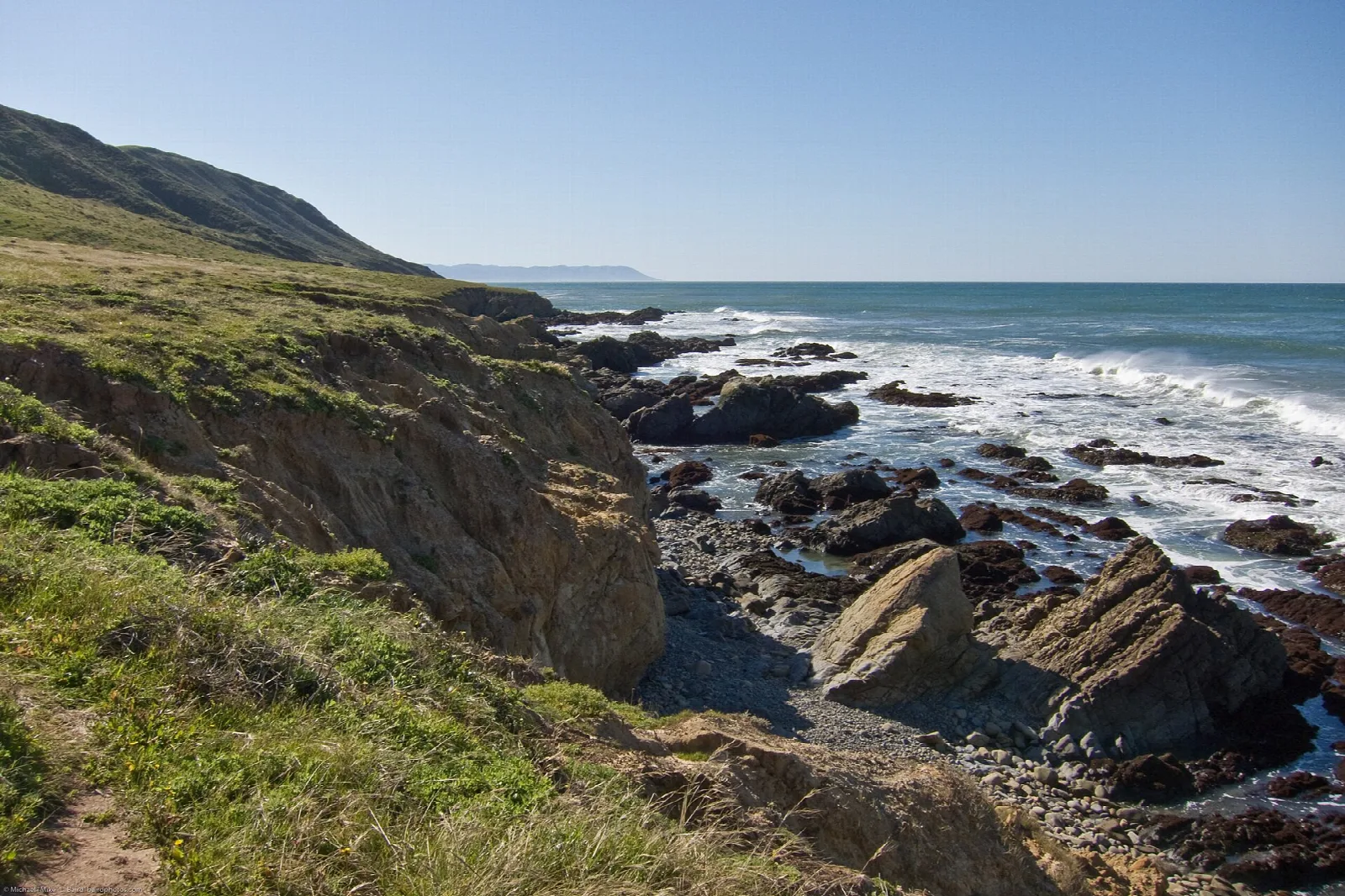 Grassy bluff and rocky shore with surf under clear sky