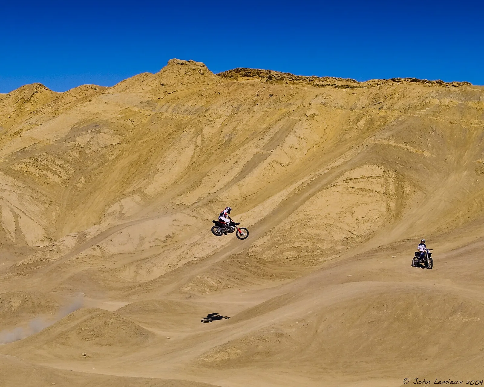 Riders on dirt bikes on a tan badland hillside under blue sky