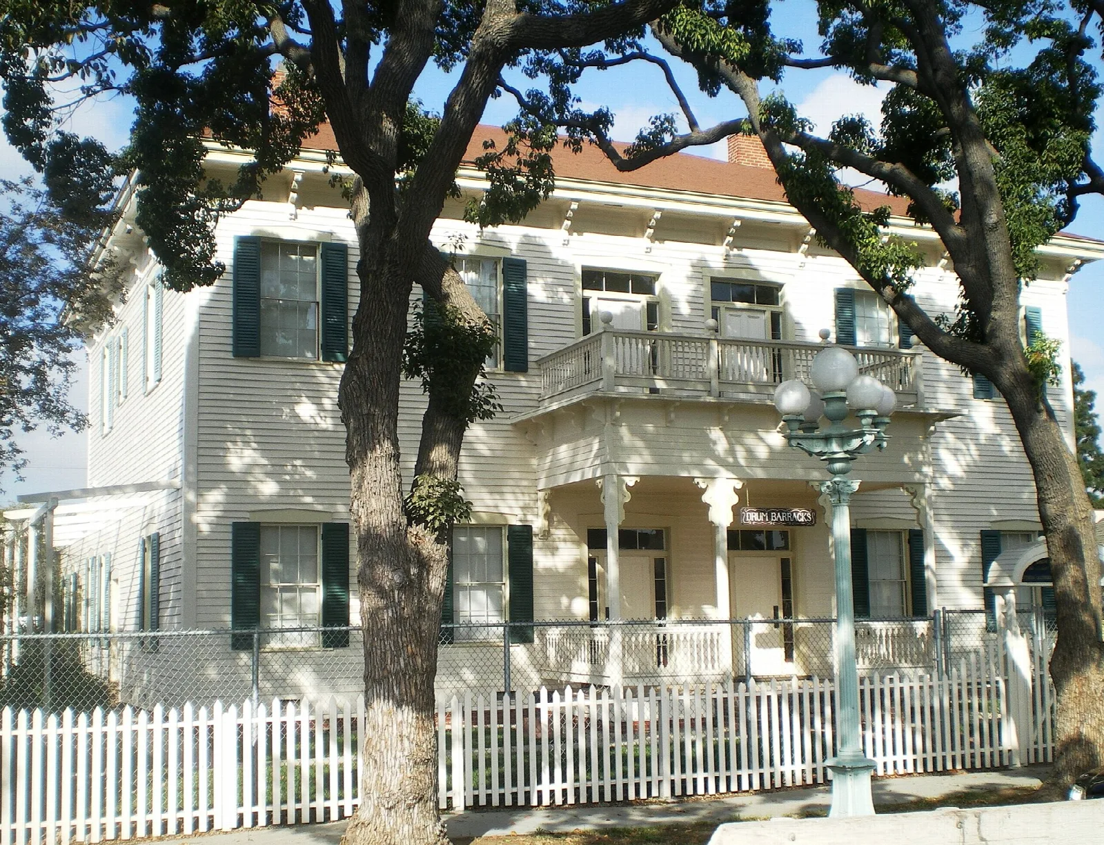 White two-story house with balconies and a picket fence under large trees