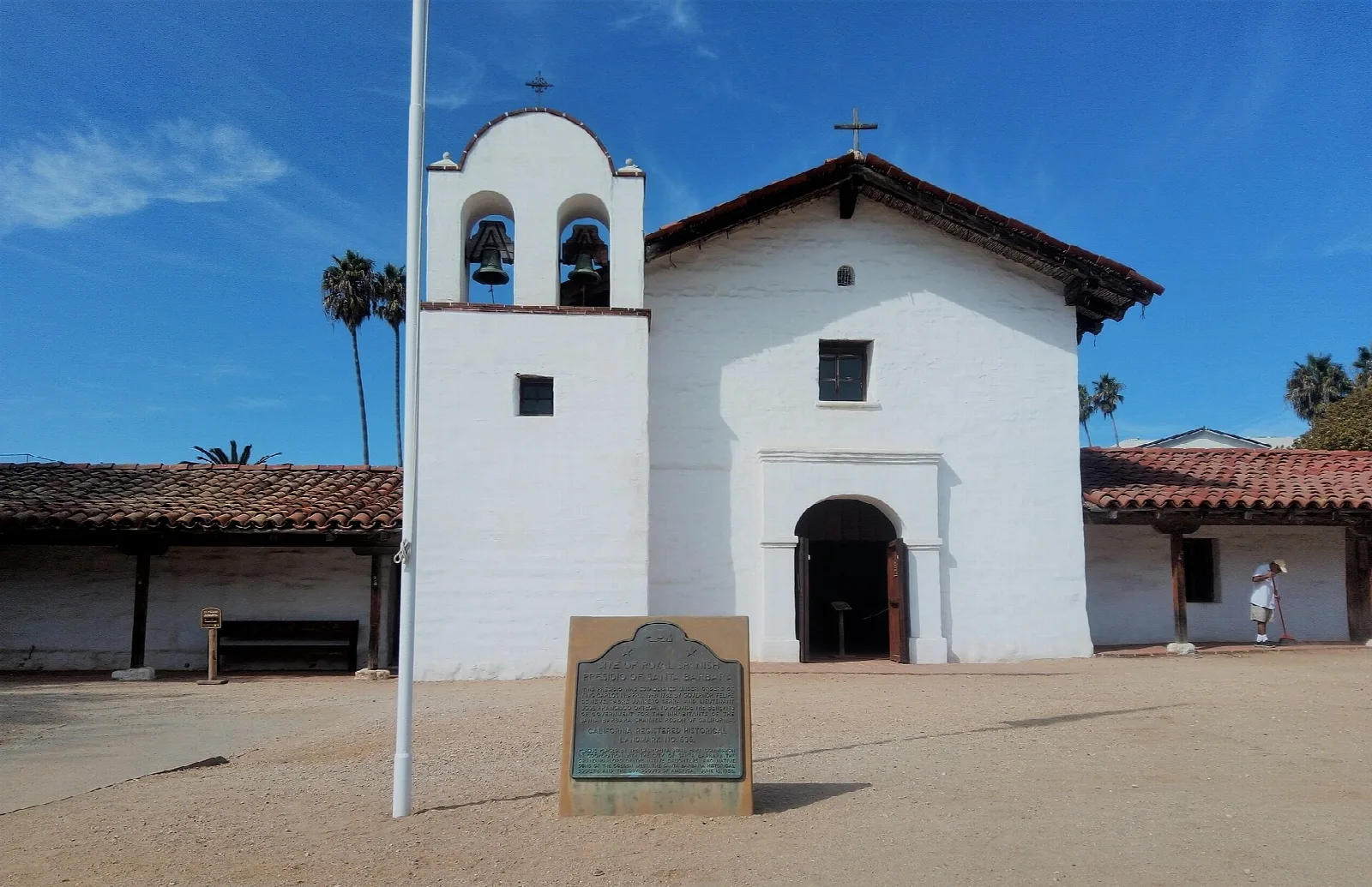 Chapel with twin bells and a plaque in front, on a sunny plaza
