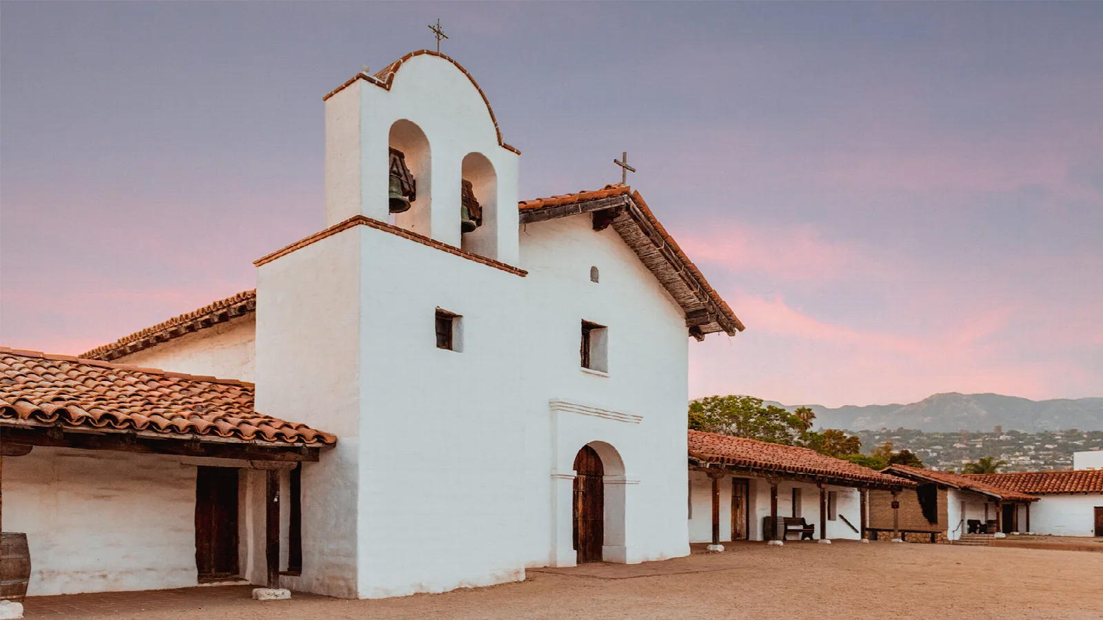 White adobe chapel with a red tile roof and bell gable under a pink evening sky