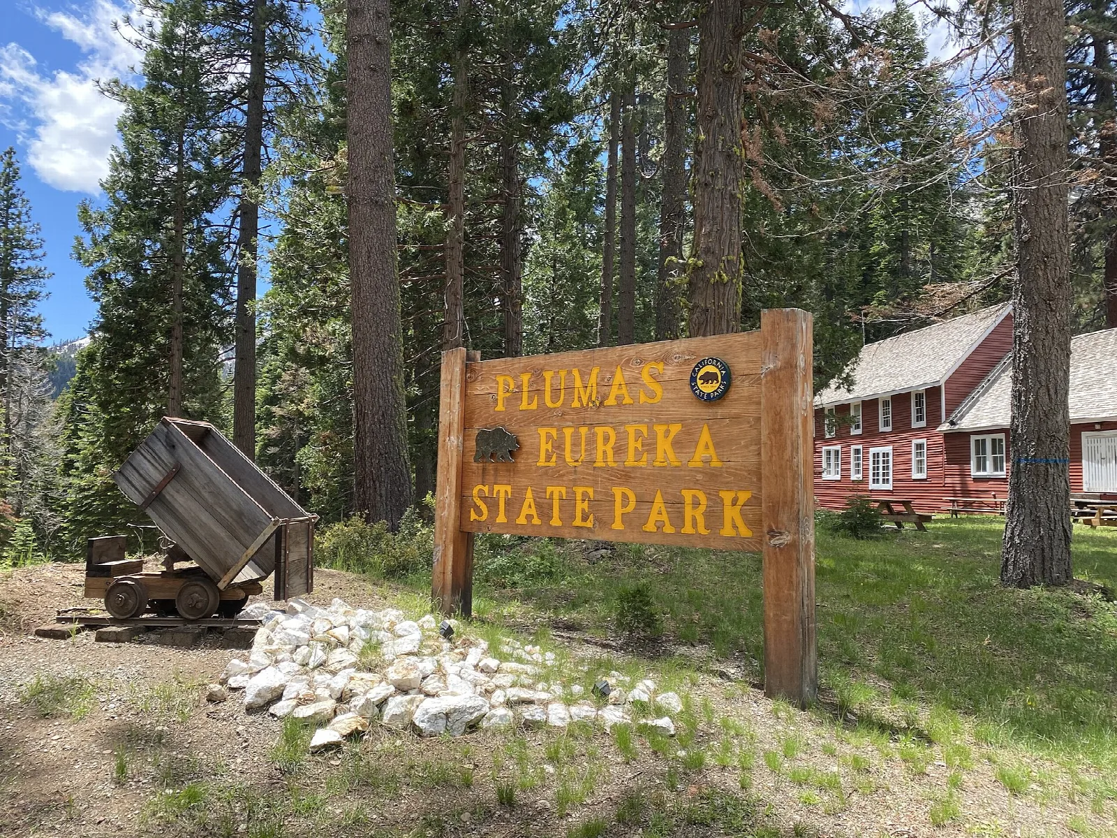 Wooden park sign in front of a red building and an old ore cart under tall pines