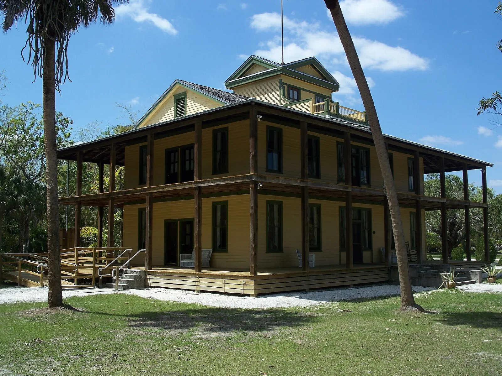 Two-story yellow wooden house with wraparound porches and palms