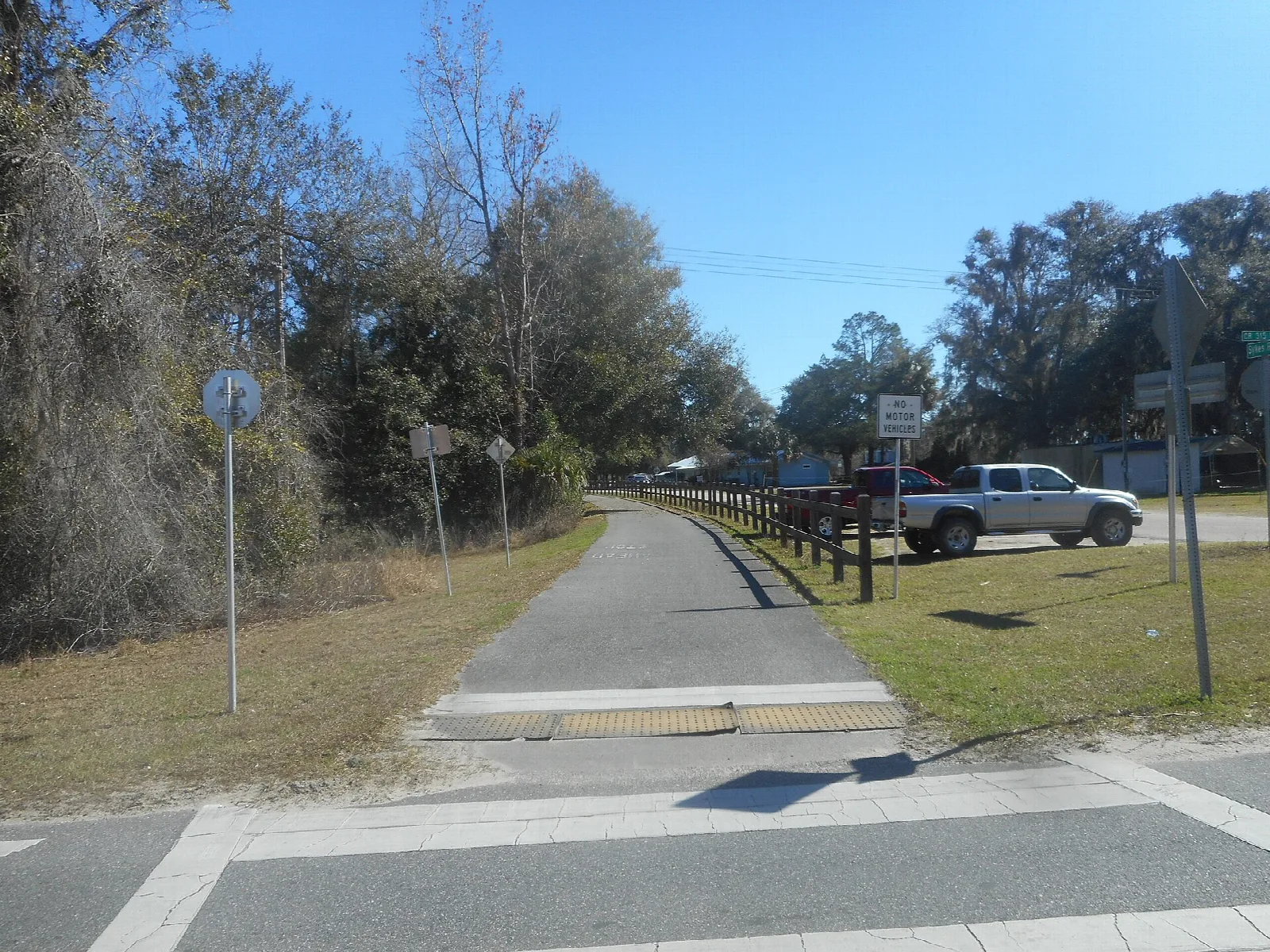 Paved trail crossing a road between trees and a fence