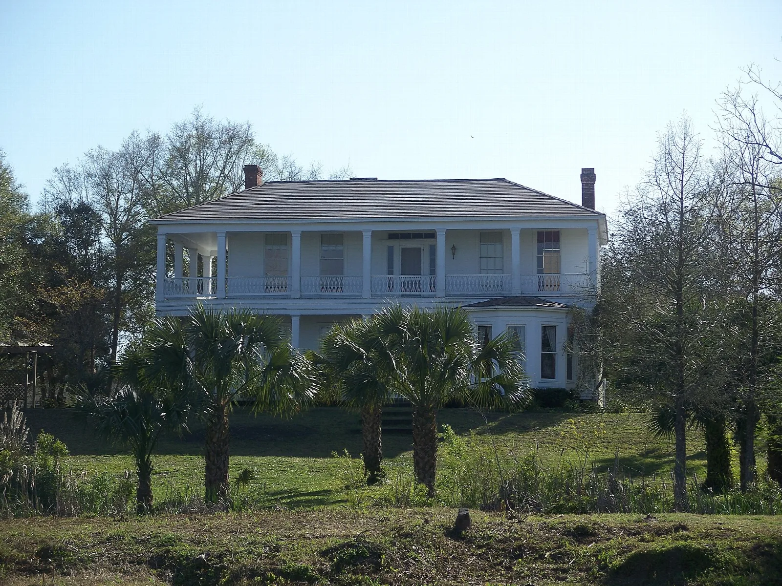 Two-story house with a long porch seen across low ground and palms