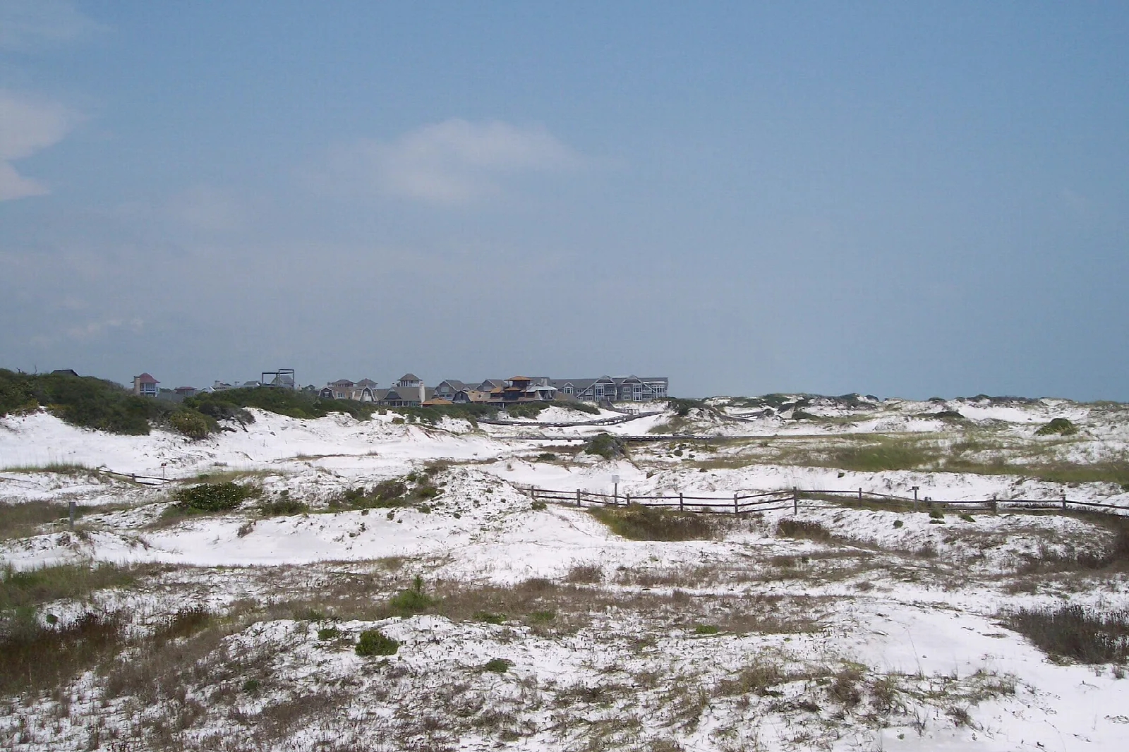 White sand dunes with sparse grass and distant buildings under blue sky