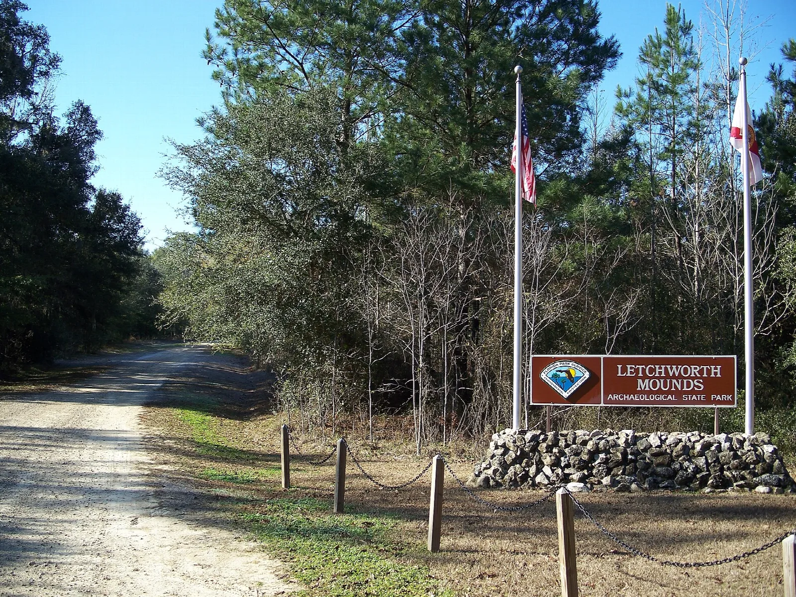 Gravel road with flags and a park sign by a low stone pile