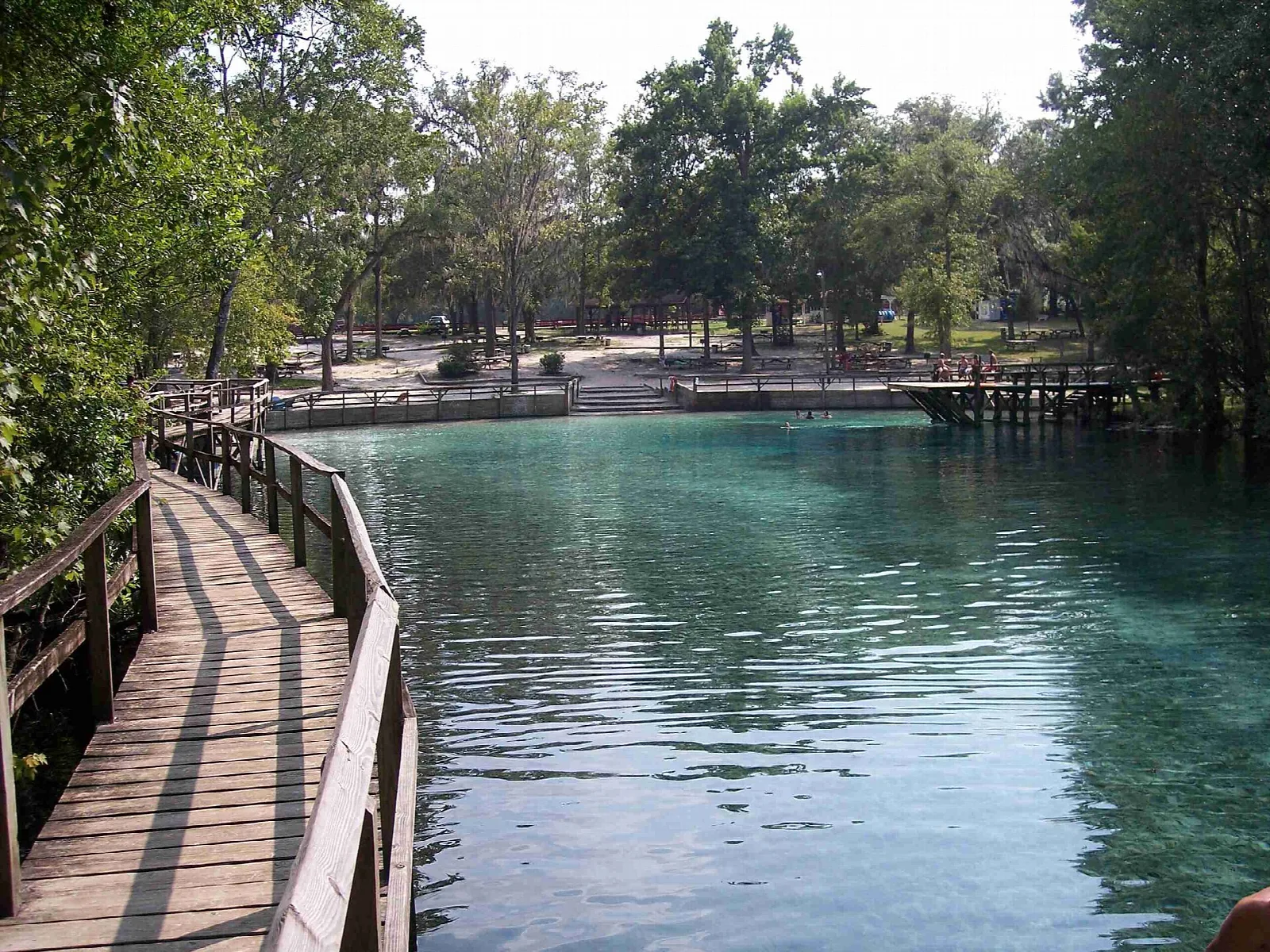 Wood boardwalk beside a clear blue-green spring with picnic area beyond