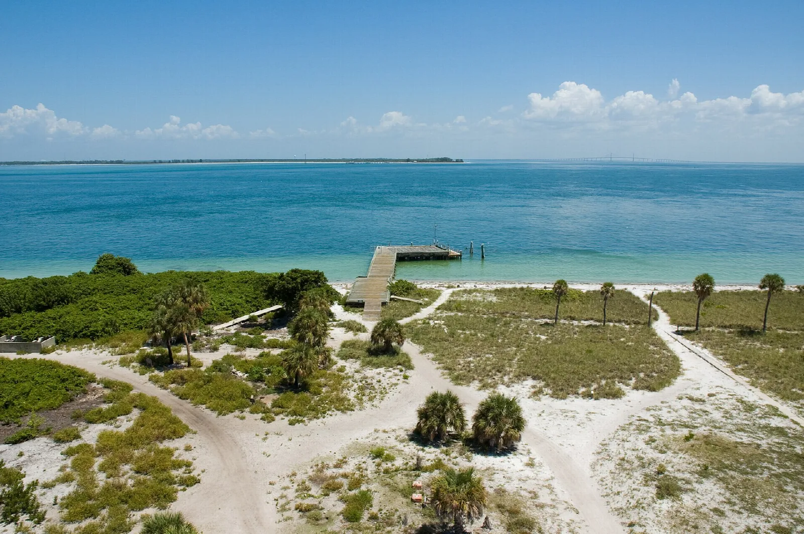 Blue water, a short pier and a sandy island with green scrub