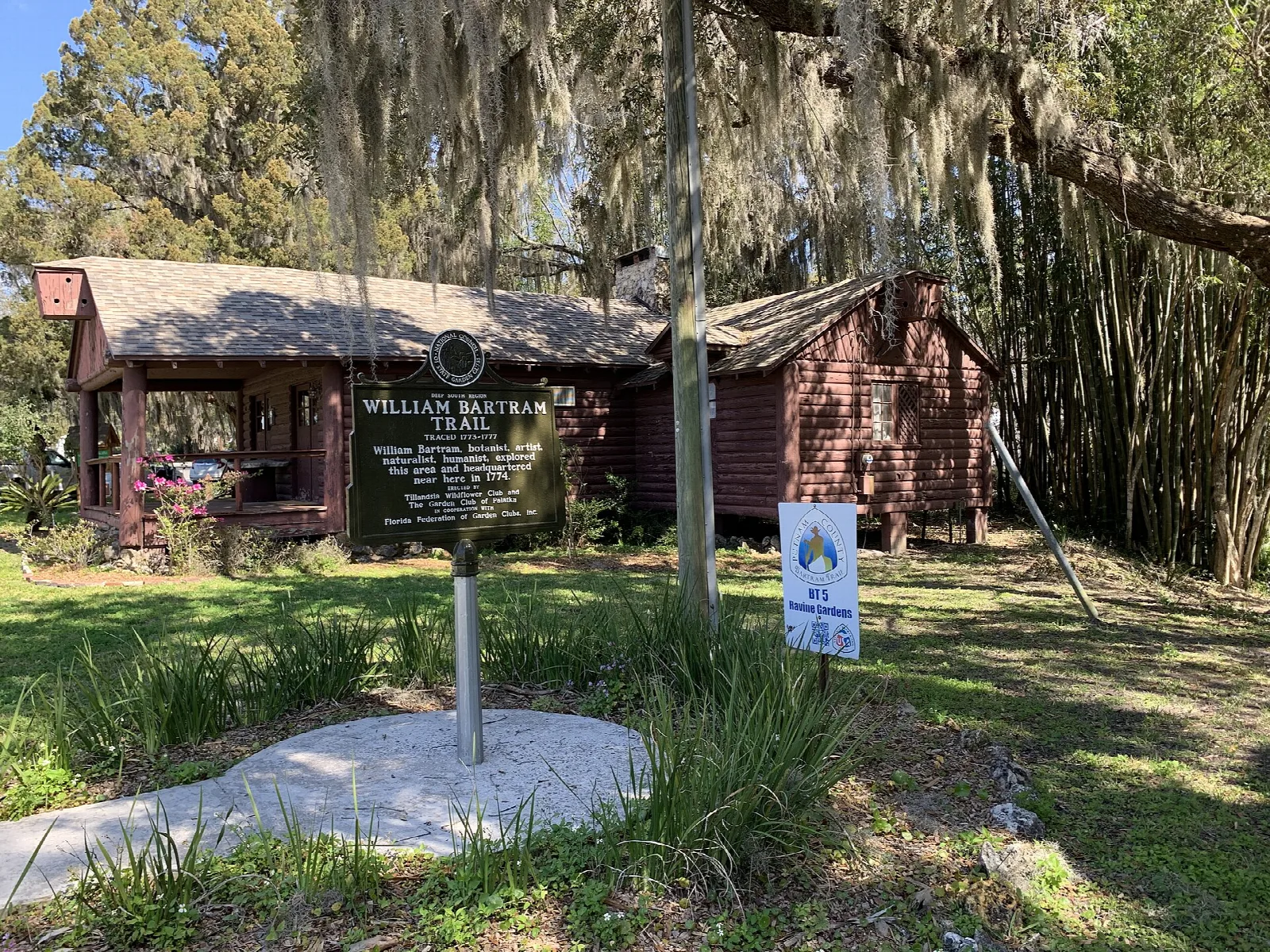 Small log-roofed shelter with a historical marker under moss-hung trees